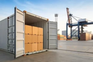 Open shipping container filled with boxes at a seaport terminal, with cranes and stacked containers in the background.
