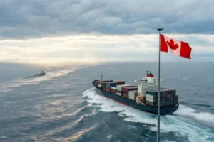 Large cargo ship carrying containers, sailing at sea with a Canadian flag flying in the foreground under a cloudy sky.