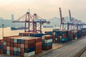 Port scene with cranes loading containers next to a road. Large ship visible on the horizon, city skyline in background.