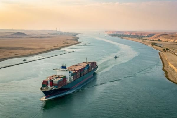 A container ship navigating through a narrow strait with geopolitical tension implied.